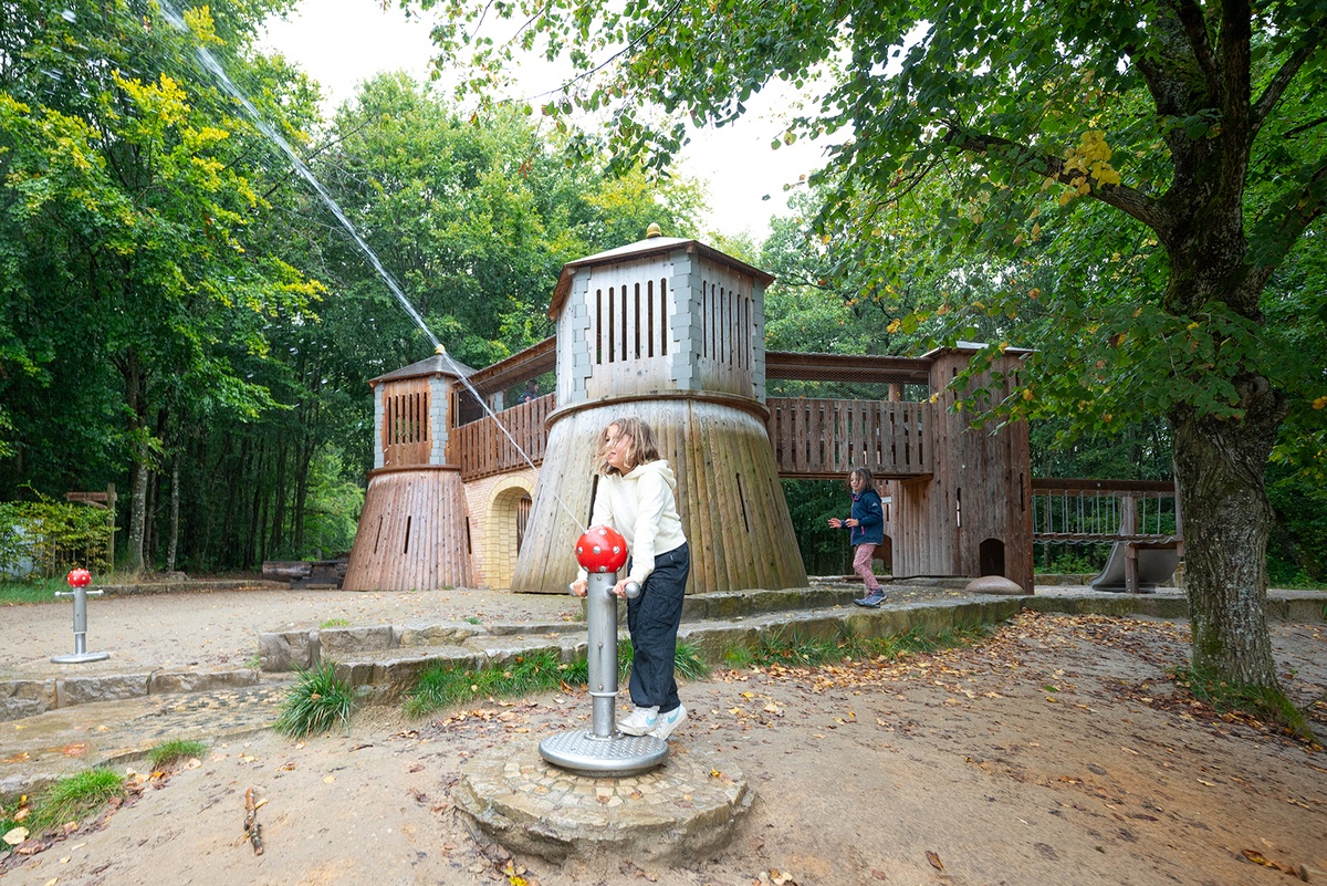 Zwei Kinder spielen auf einem Spielplatz mit einer Burganlage aus Holz im Hintergrund. Eines der Kinder bedient eine Wasserspritzpumpe, während das andere Kind am Eingang der Burg steht. Der Spielplatz ist von Bäumen umgeben.