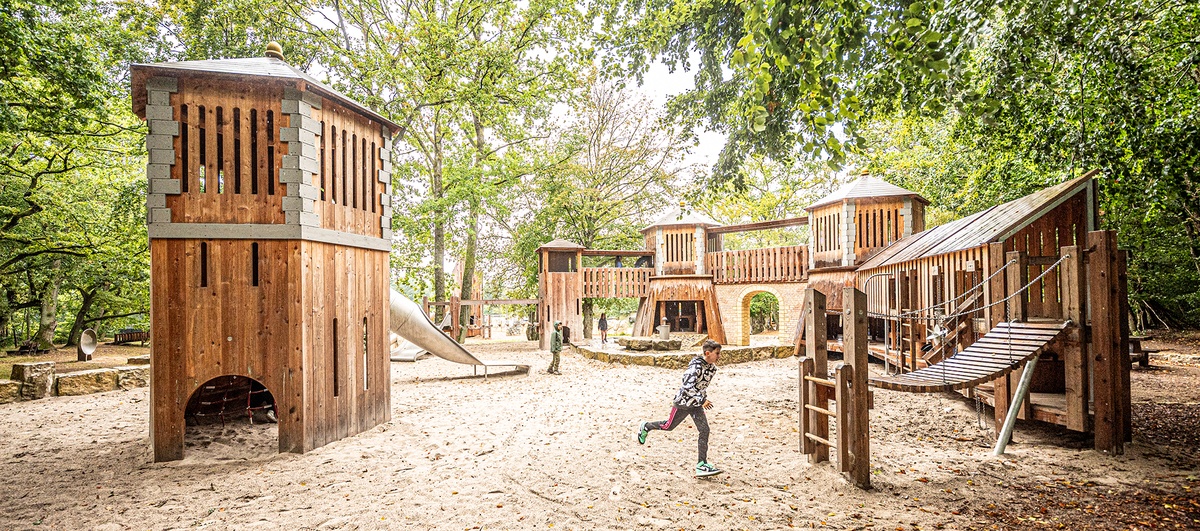 Mehrere Kinder spielen auf einem Spielplatz mit Burganlage aus Holz. Ein Teil der Tunnelrutsche ein Aufgang zu einer kleinen Hängebrücke aus Holz ist zu sehen. Der Spielplatz ist von Bäumen umgeben und liegt in einer natürlichen, waldähnlichen Umgebu