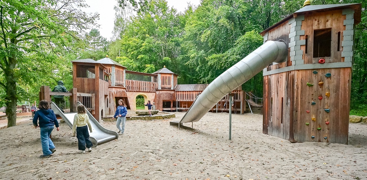Mehrere Kinder spielen auf einem Spielplatz mit Burganlage aus Holz. Im Vordergrund ist ein Turm mit Klettergriffen und Tunnelrutsche und eine breite Kleinkindrutsche zu sehen. Der Spielplatz ist von Bäumen umgeben.