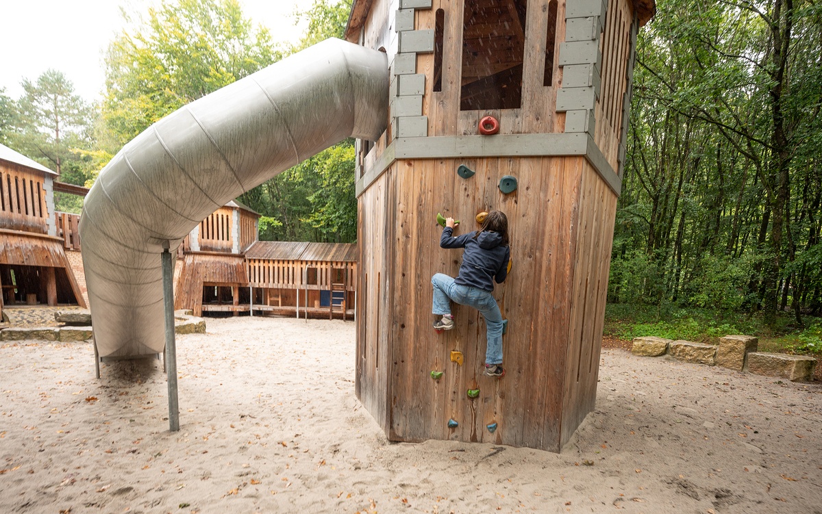 Ein Junge in Jeans und dunkelblauem Pullover klettert die Kletterwand eines Holzturm hoch, an der seitlich eine Tunnelrutsche nach unten führt. Im Hintergrund ist eine Burganlage und Spielwand aus Holz zu sehen. Der Boden ist sandig.