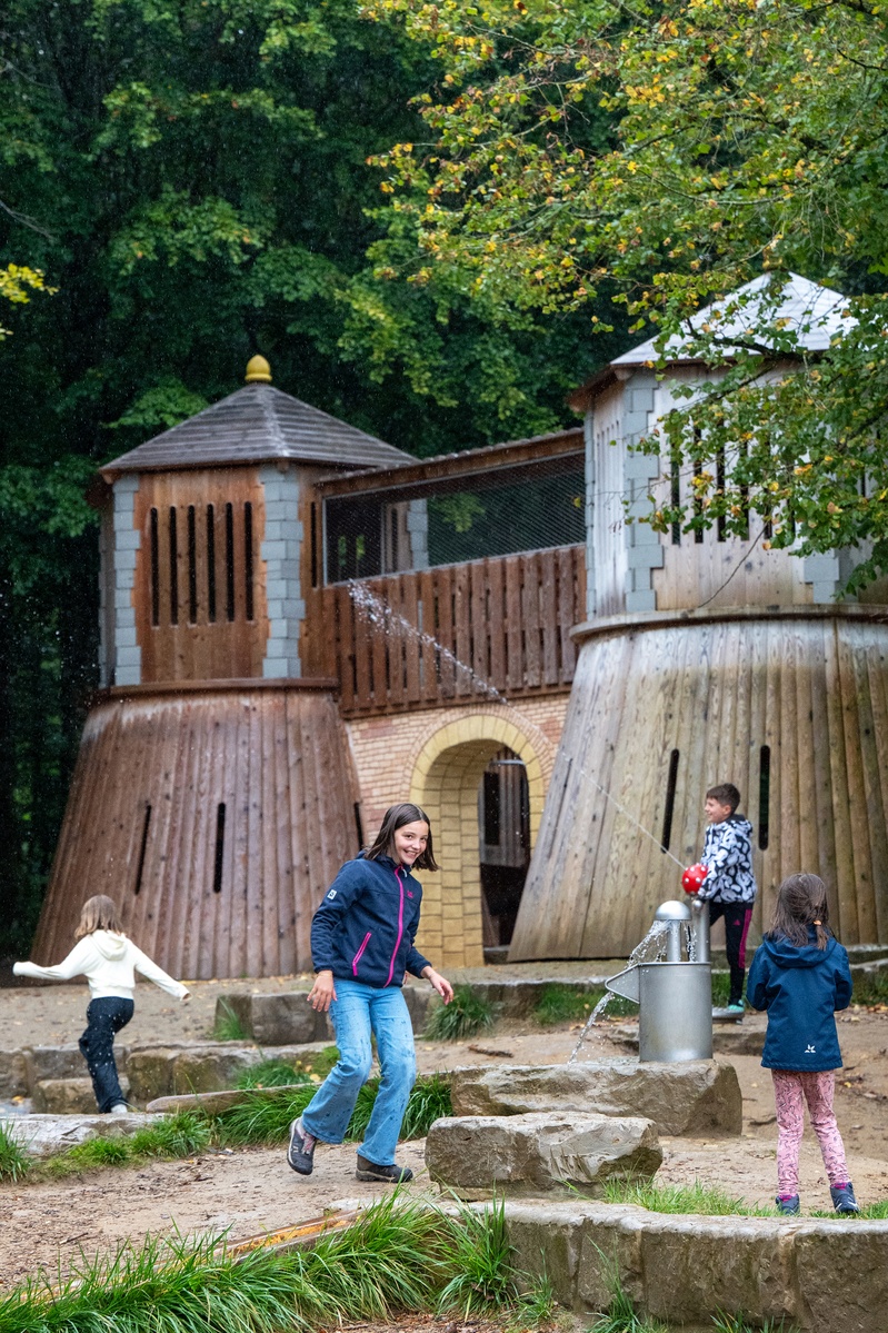 Mehrere Kinder spielen auf einem Spielplatz mit einer Burganlage aus Holz im Hintergrund. Eines der Kinder bedient eine Wasserspritzpumpe und aus dem Wasserspender davor läuft Wasser. Der Spielplatz ist von Bäumen umgeben.