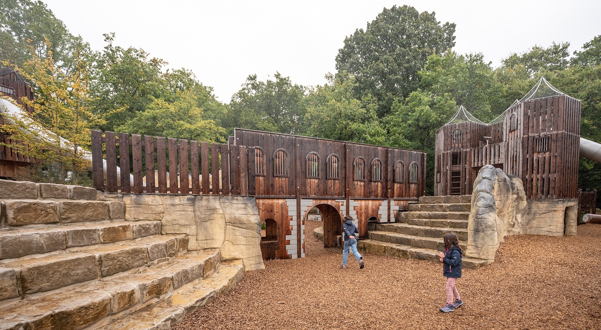 Ein Spielplatz mit einer Burganlage aus Holz, die mehrere Ebenen und bogenförmige Durchgänge hat. Links führen Steinstufen zur Anlage hinauf. Der Boden gleicht einem Waldboden, im Hintergrund stehen Bäume. Zwei Kinder sind im Vordergrund zu sehen.