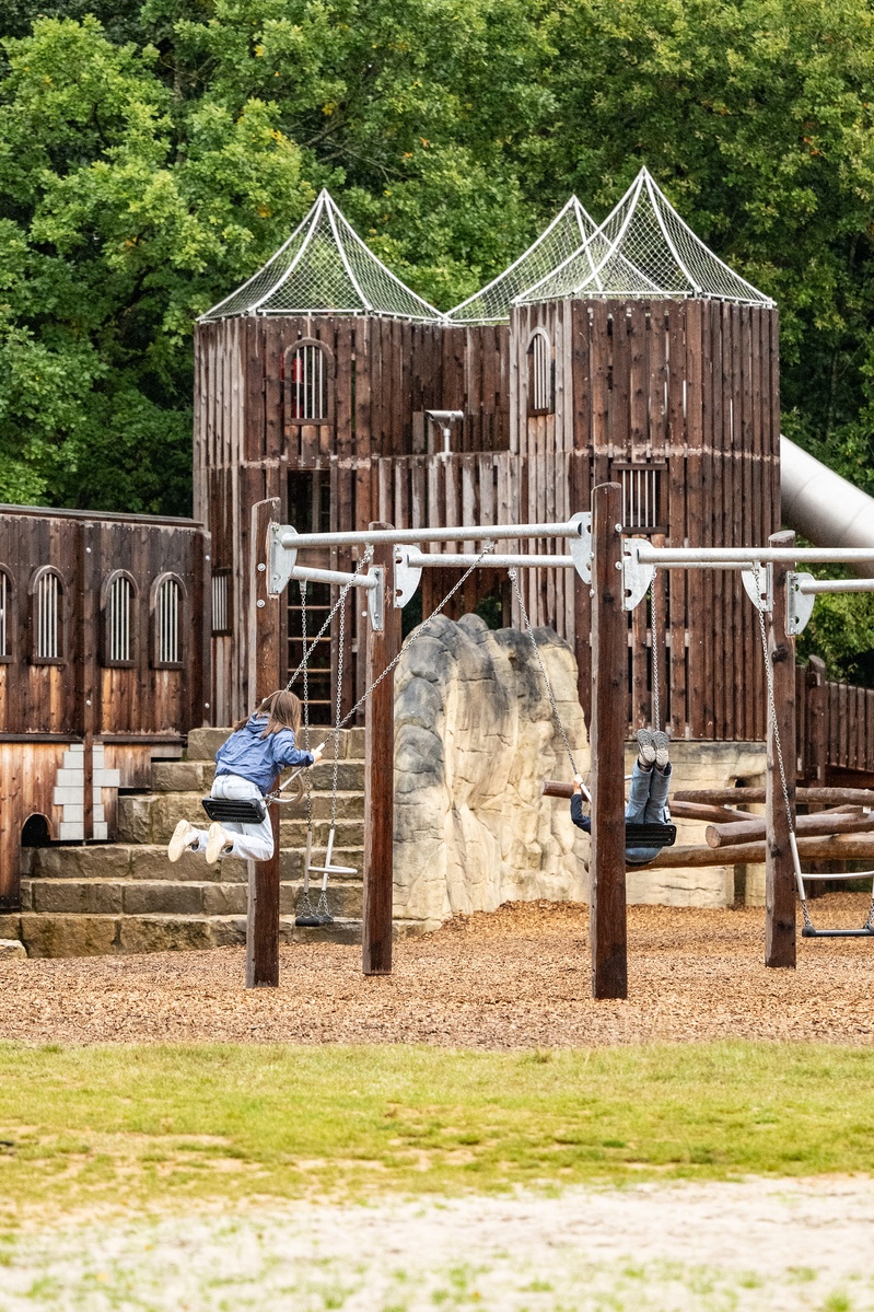 Zwei Kinder schaukeln auf einer Sechseckschaukel. Im Hintergrund ist eine Burganlage aus Holz und ein Teil der Tunnelrutsche zu sehen. Der Spielplatz ist von Bäumen umgeben und liegt in einer natürlichen, waldähnlichen Umgebung.