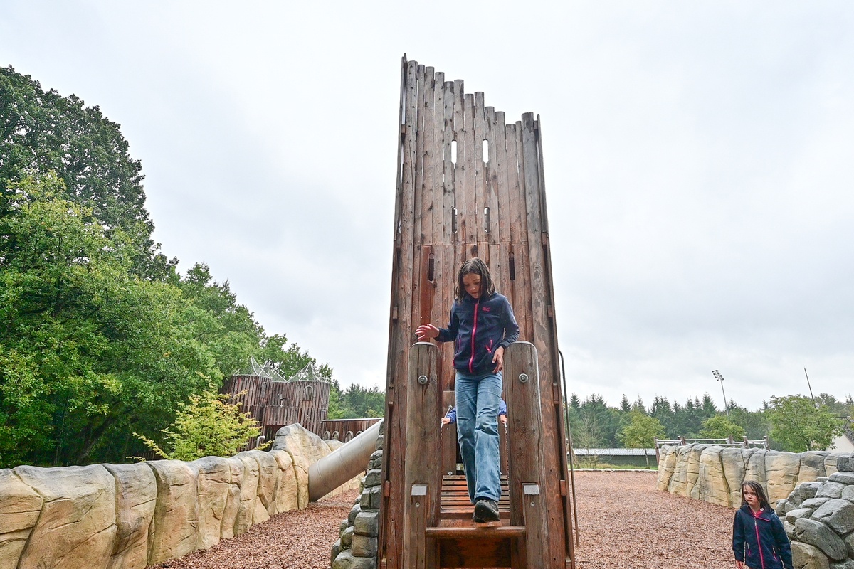 Ein Spielplatz mit oben abgeschrägtem Palisadenturm aus Holz. Ein Mädchen überquert die Hängebrücke. Der Spielplatz ist von Bäumen umgeben und hat ein natürliches, rustikales Design mit steinähnlichen Abgrenzungen und Holzschnitzeln auf dem Boden.