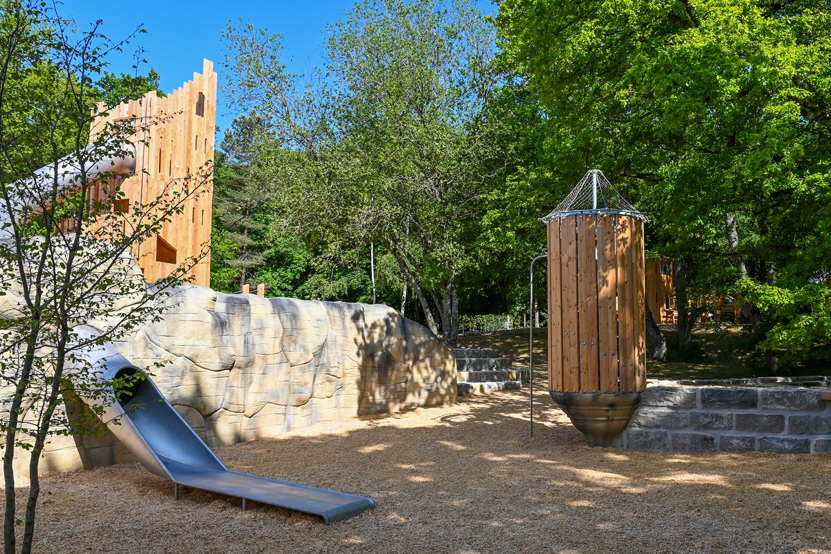 Ein Spielplatz mit abgeschrägtem Holzturm und Tunnelrutsche. Rechts steht eine zylindrische Holzkonstruktion mit einem konischen Dach und Rutschstange. Im Hintergrund sind Bäume und Grünflächen zu sehen, die Schatten spenden.