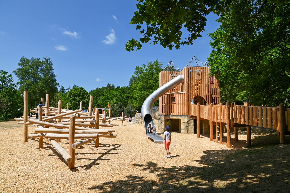 Ein Außen-Spielplatz mit Kletterstruktur und Burganlage mit Tunnelrutsche aus Holz. Mehrere Kinder spielen auf dem Gelände. Der Boden ist mit Holzschnitzeln bedeckt, im Hintergrund sind Bäume zu sehen. Der Himmel ist wolkenlos.