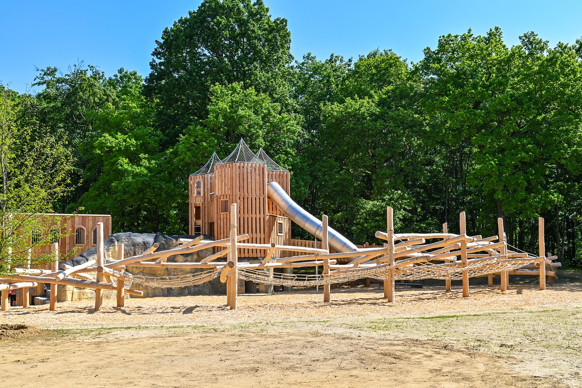 Spielplatz mit großer Kletterstruktur im Vordergrund. Im Hintergrund ist eine Burganlage aus Holz mit Tunnelrutsche zu sehen. Der Spielplatz ist von Bäumen umgeben, der Himmel ist blau und wolkenlos.