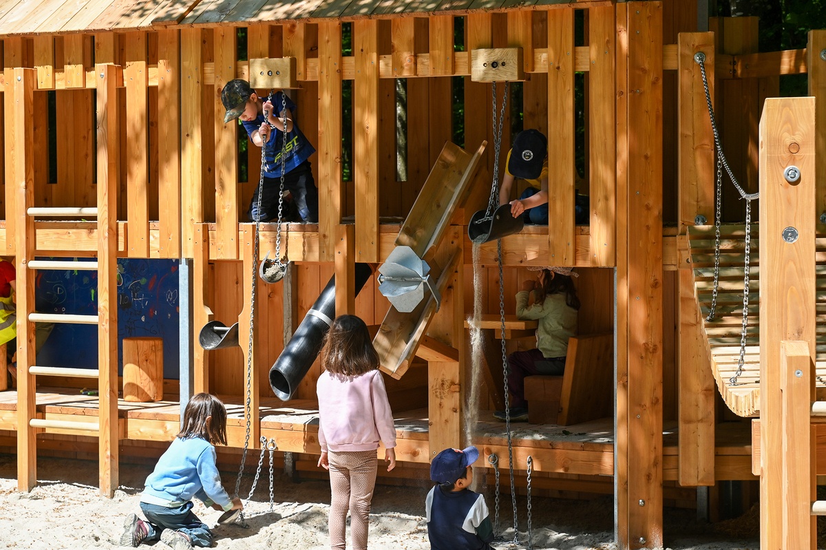 Mehrere Kinder spielen an einer Spielwand aus Holz mit Sandspieleinrichtungen wie Sandaufzug, Rinnen und Sandrad.  Auf der rechten Seite ist ein Teil der Hängebrücke zu sehen.