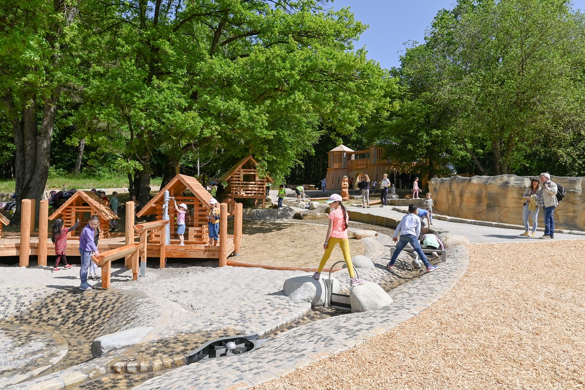 Außenspielanlage mit kleinen Spielhäusern aus Holz und Wasserspielplatz. Kinder spielen mit den Wasserspielgeräten, während Erwachsene sie beaufsichtigen. Der Spielplatz liegt in einem parkähnlichen, von Bäumen umgebenen Gelände.