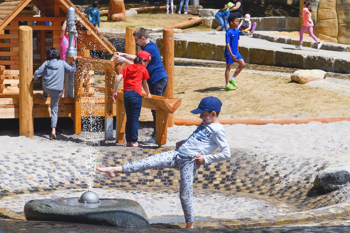 Ein lachender Junge mit blauem Cap spielt mit einer Wasserspritzdüse. Dahinter ist ein Spielhaus aus Holz mit Holzrinnen und einer Spielplatzpumpe zu sehen, an der mehrere Kinder spielen. Im Hintergrund sitzen oder gehen weitere Kinder.