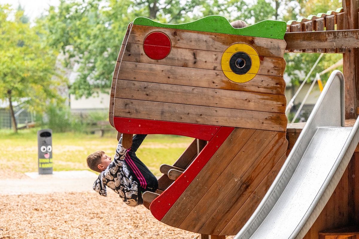 Kinder spielen auf der Plattform Drache "Fauchi" mit Rutsche und Kletterstange. Im Hintergrund sind Sonnensegel zu sehen, die an hohen Holzpfosten befestigt sind. Der Himmel ist hell, aber bewölkt.