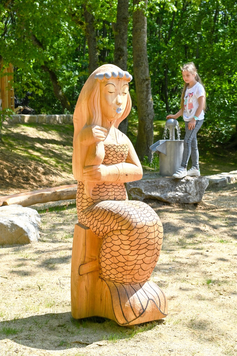 Skulptur einer sitzenden Meerjungfrau mit geschlossenen Augen, aus Holz gefertigt. Dahinter betätigt ein Mädchen einen Wasserspender. Im Hintergrund sind Bäume zu sehen, die Schatten spenden.