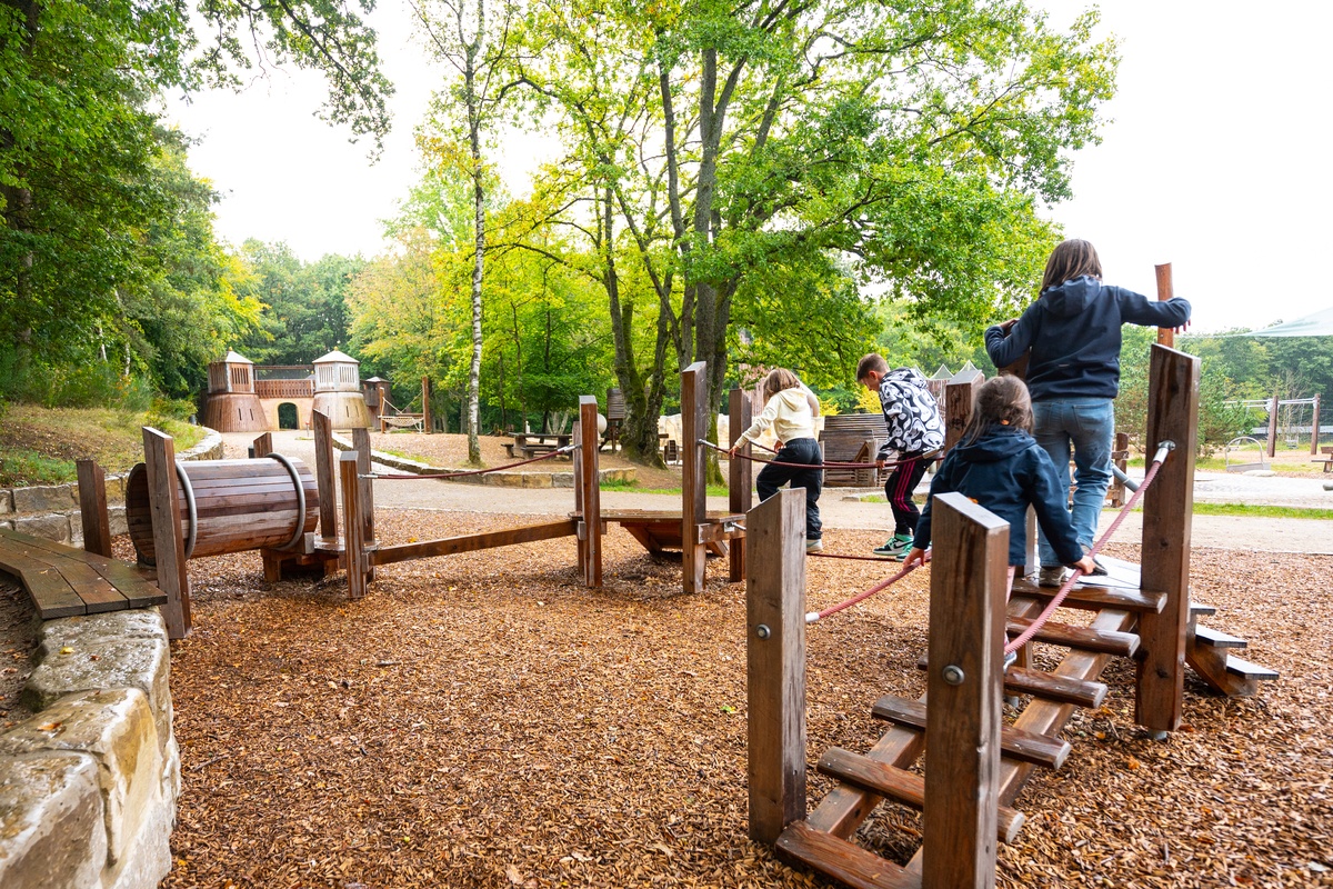 Kinder spielen auf einem Kleinkindparcours, der sich auf einem Außenspielplatz befindet und von Bäumen umgeben ist. Zwei Kinder steigen einen Holzsteg mit Seilhandläufen nach oben, zwei andere Kinder überqueren ein Balancierseil mit Halteseilen.