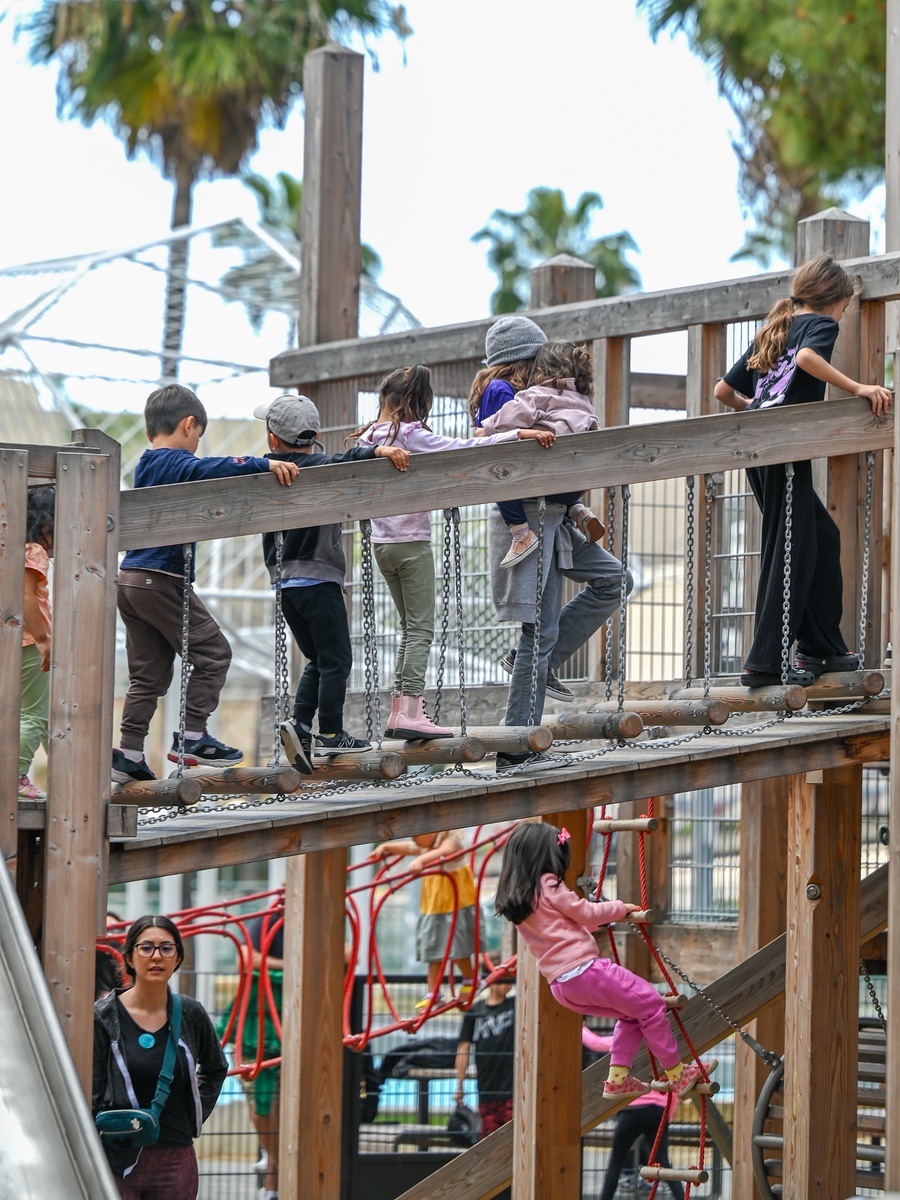 Kinder auf Hängebrücke mit Handlauf und Laufhölzern, die zur Spielpyramide hinauf führt. Dahinter ein Mädchen auf Strickleiter © Daniel Perales