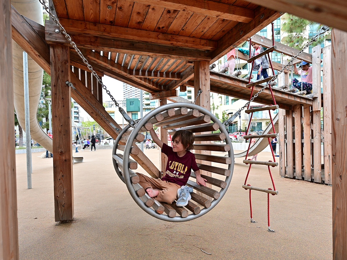 Ein Junge sitzt in einem an Ketten aufgehängten Kriechfass aus Holz, das sich unter der Spielpyramide befindet. Daneben sieht man eine Strickleiter, einen Brückenübergang mit Kindern darauf und die Unterseite der Tunnelrutsche © Daniel Perales