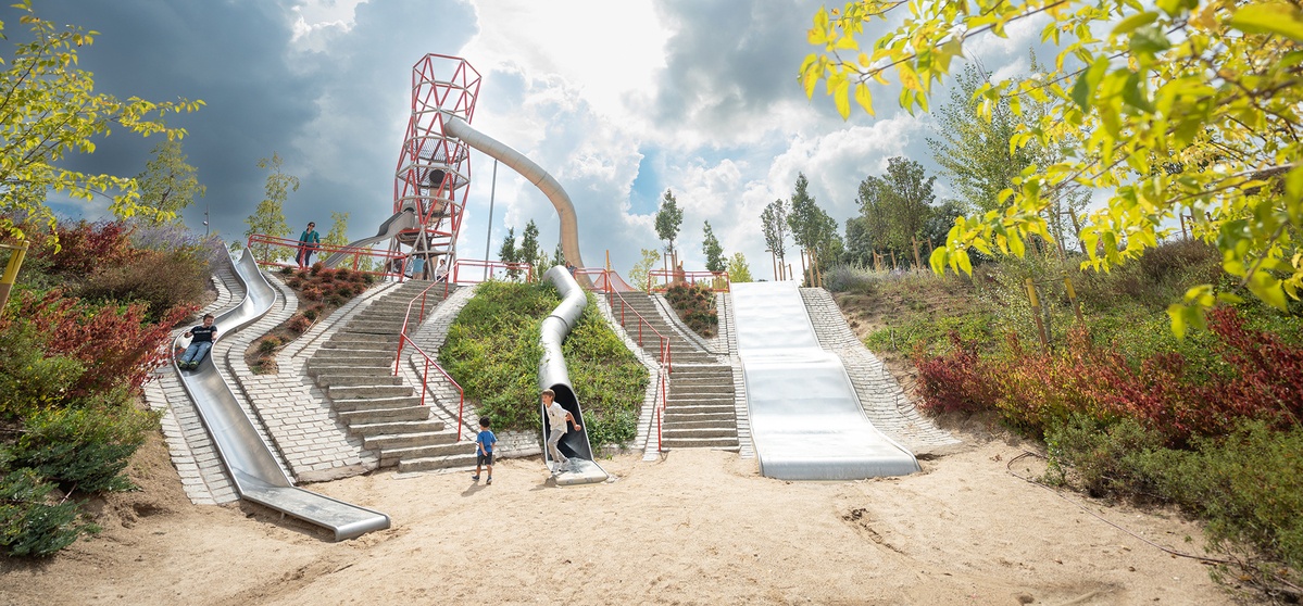 Play area with several slides and a special stainless steel tower. The slides are built into a hill with stairs leading upwards. The play area is surrounded by trees and bushes, the sky is partly covered with dark clouds © Daniel Perales