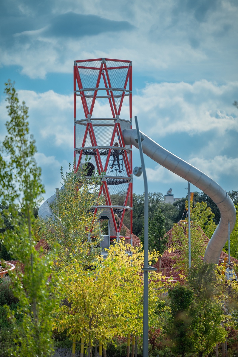 A special stainless steel tower surrounded by bushes and trees with a tunnel slide. A child climbs up the net on the tower.  The sky is partly cloudy © Daniel Perales