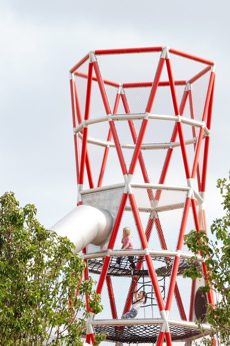 A special tower made of stainless steel and red support elements. Two girls climb up the nets in the tower. The entrance to the tunnel slide on the tower can be seen. There are trees and bushes in the front © Daniel Perales