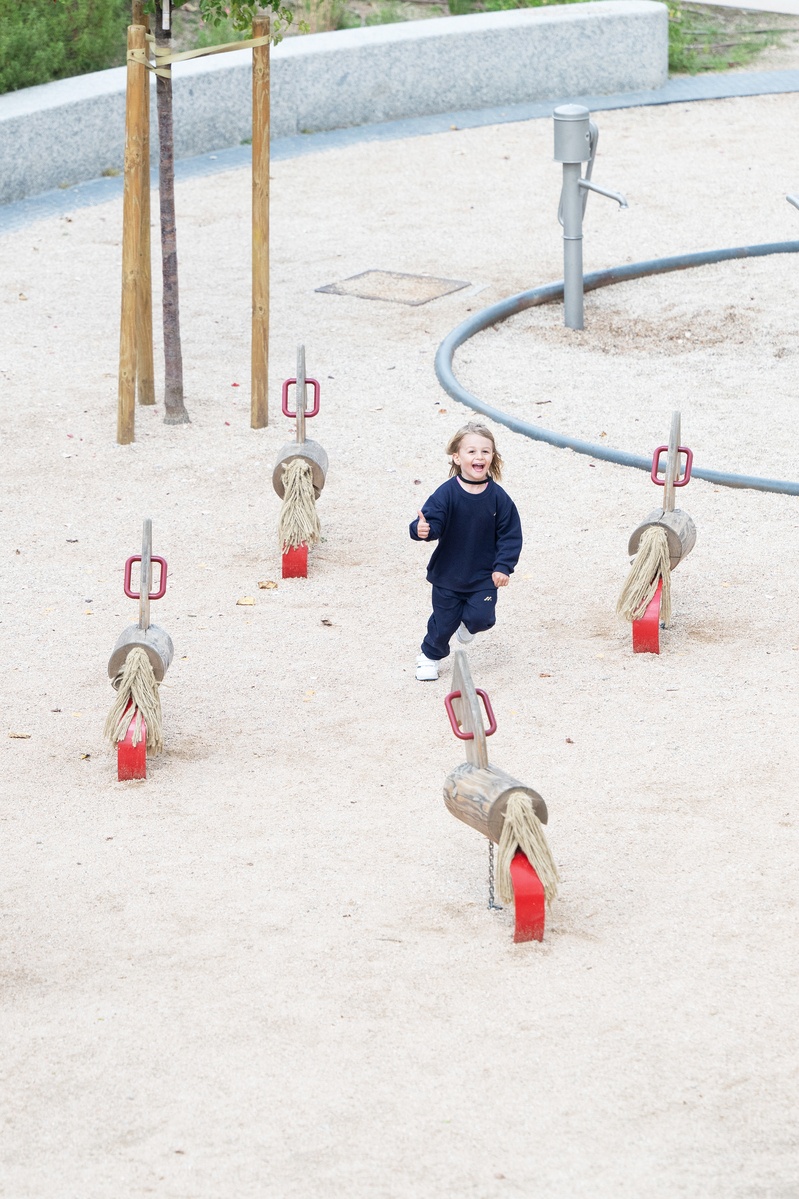 A laughing girl in dark blue clothes walks on sandy ground. Surrounding her are four wooden swinging horses. In the background are parts of a tree and a stone wall and an unused stainless steel playground pump © Daniel Perales