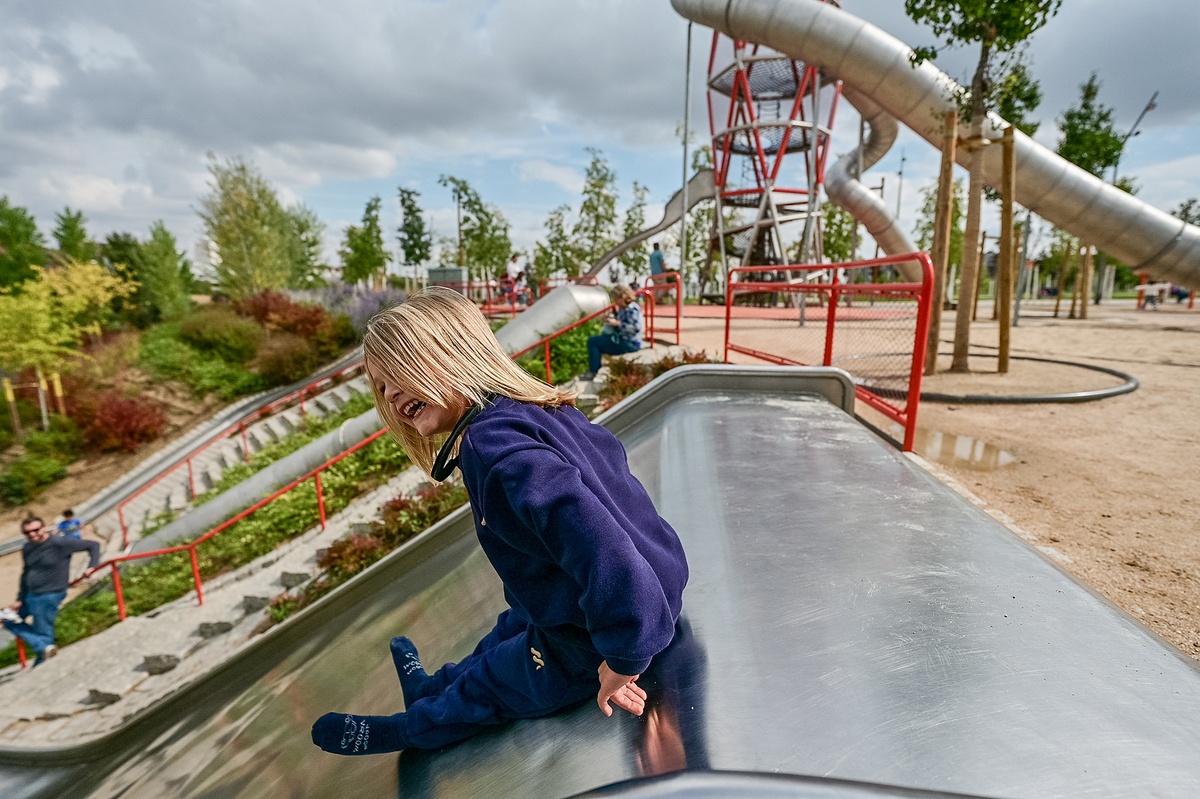 Ein blondes Kind sitzt lachend am oberen Ende einer flachen breiten Rutsche. Dahinter sieht man eine buschige Landschaft, Teile der Tunnelrutsche und des Sonderturms aus Edelstahl © Daniel Perales