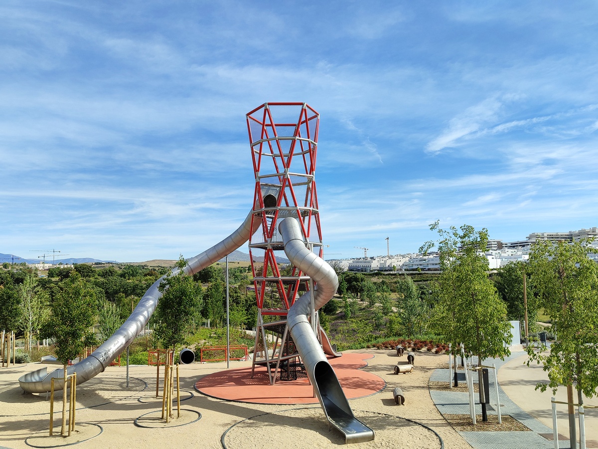 Special stainless steel tower with tunnel slides and several wooden sheeps standing and lying on the ground next to it. In the background, a landscape of trees and a white-blue sky © Daniel Perales