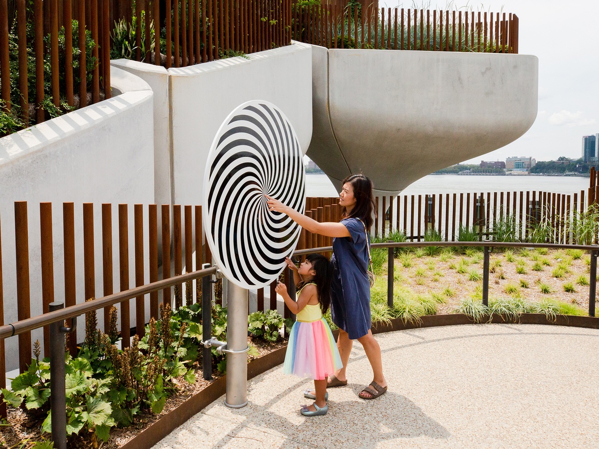 A mother and her daughter move a rotating disc mounted on a steel post. A concrete structure, green areas, water and buildings can be seen in the background.