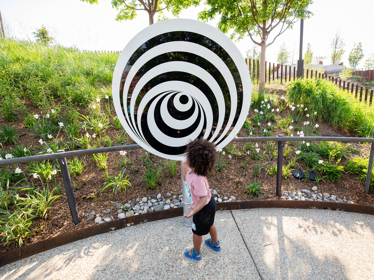 A little girl moves the rotating disc on a steel post. There is a planted hill with trees providing shade in the background. The sun is shining.