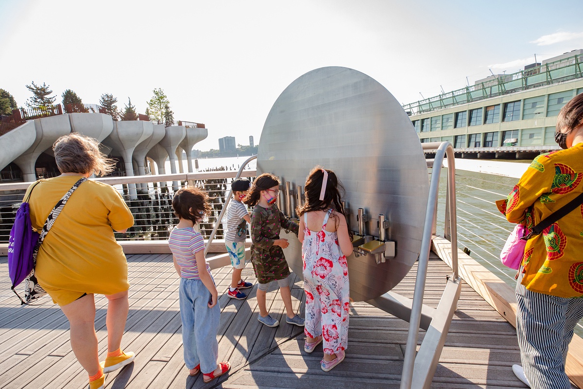 A group of people, including children and adults, are standing on a wooden deck by the water. They are gathered around the playground equipment called ‘instrument’. A modern building and architectural structures.