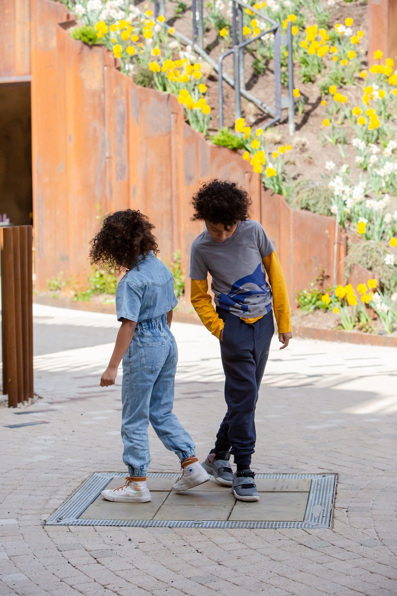 Two children bounce on the dance chimes. A hillside with yellow flowers and steel railings can be seen in the background. 