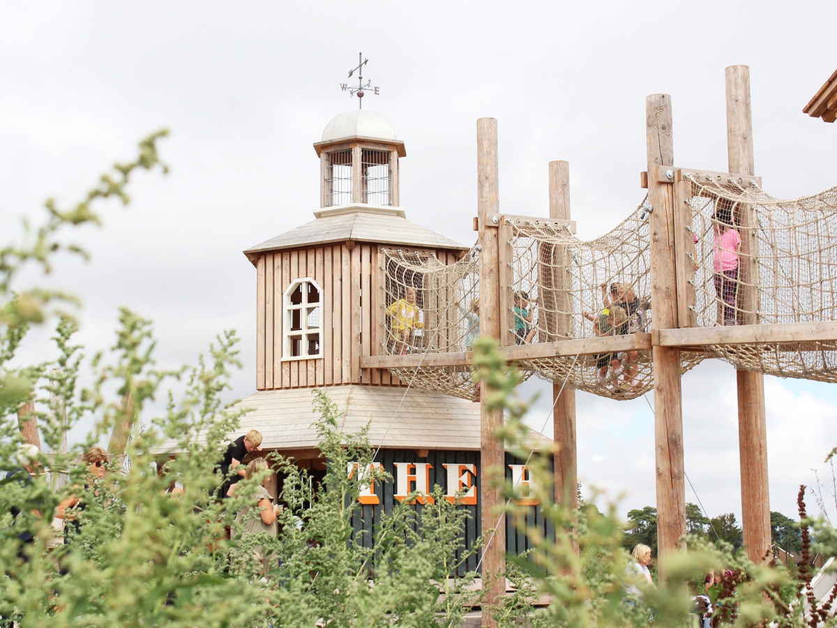 Children play on the special platform ‘The Whistling Witch’. There is a weather vane on the tower and windows on the sides. Other children are crossing a net tunnel made of natural-coloured ropes. Bushes and trees can be seen in the foreground.