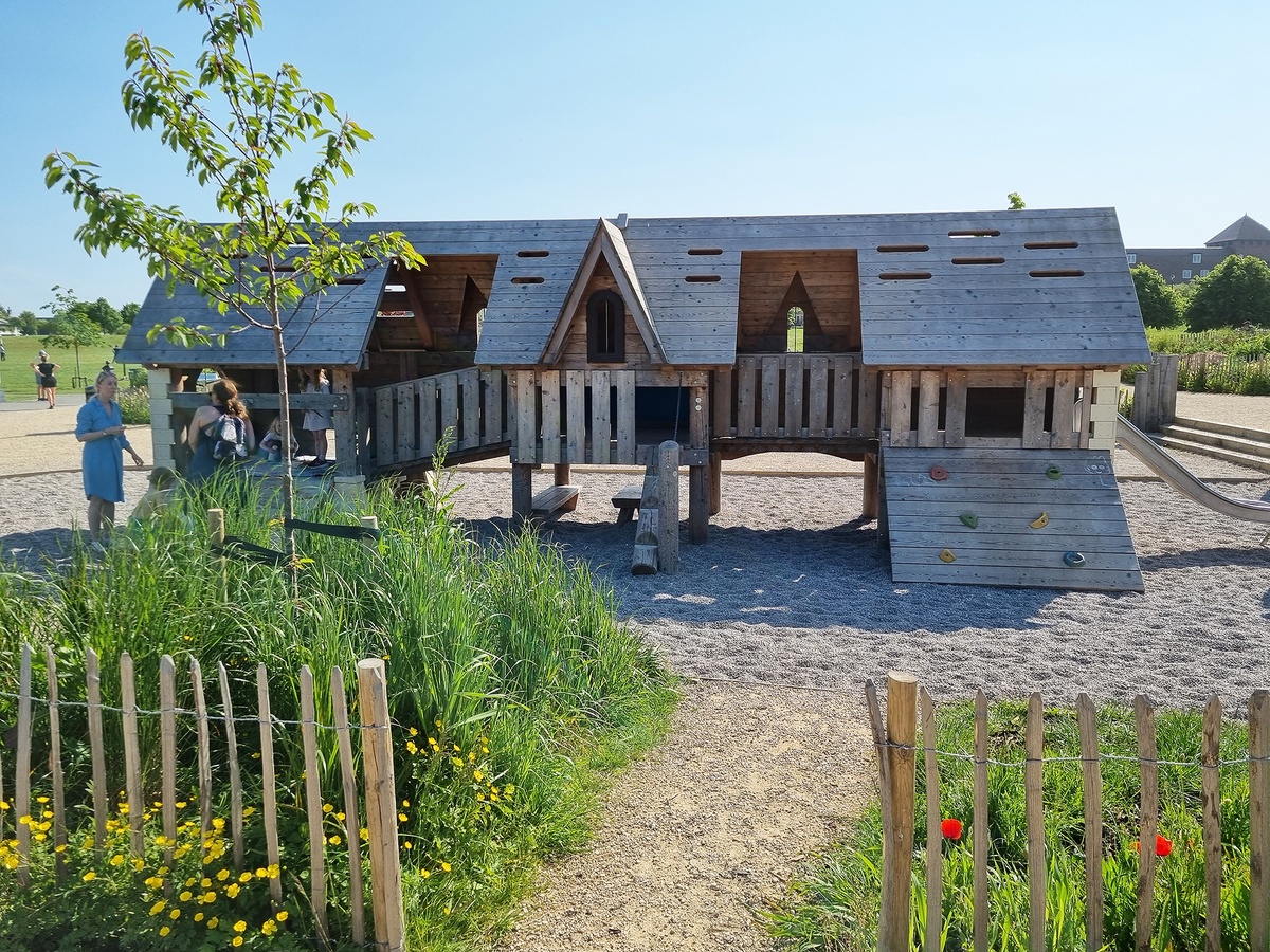 A playground with a wooden structure designed like a row of small houses with pointed roofs and various openings. Two adults are standing to the left of the houses, a child is sitting in one of the houses.