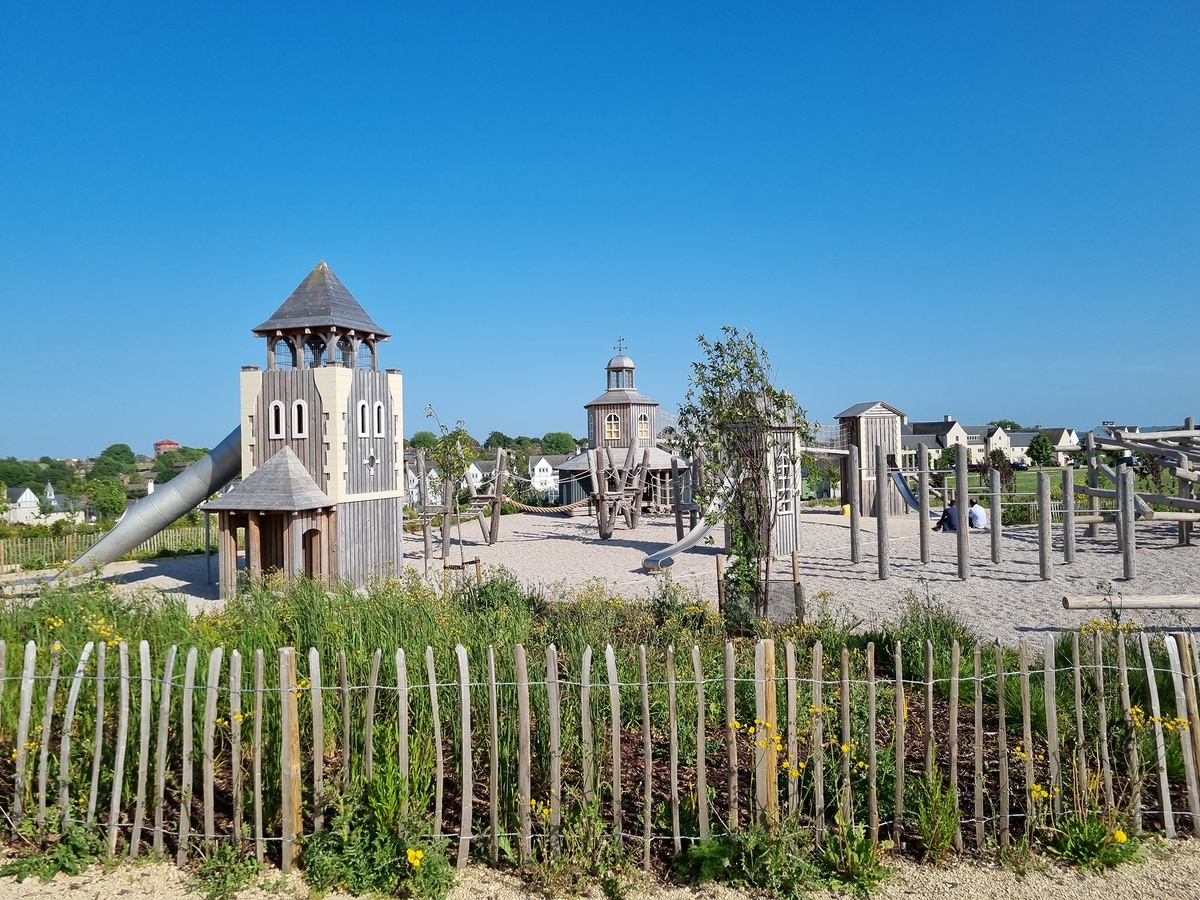 A playground with several play towers, wooden platforms, slides and elements for balancing and climbing.  The playground is surrounded by a wooden fence and lots of greenery. In the background are houses and trees under a clear blue sky.