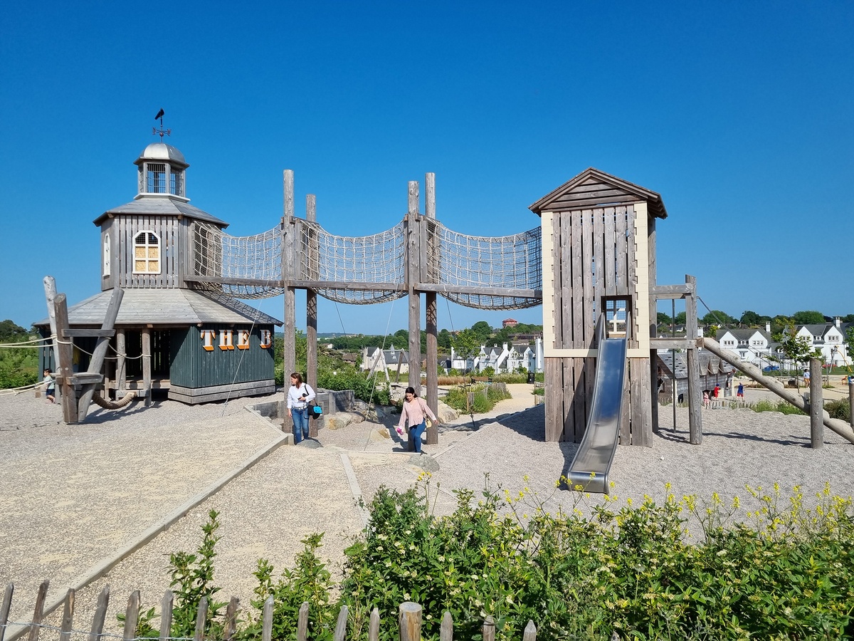 A playground with several play towers, one of which has a slide. There are wooden platforms and also elements for balancing and climbing. Two women are walking up a staircase underneath a net tunnel. The ground is covered with gravel and sand.