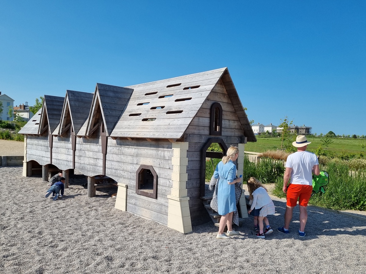 A playground with a wooden structure designed like several connected small houses. The structure has windows, doors and roofs with cut-out shapes. Three small children and two adults are playing around the structure.
