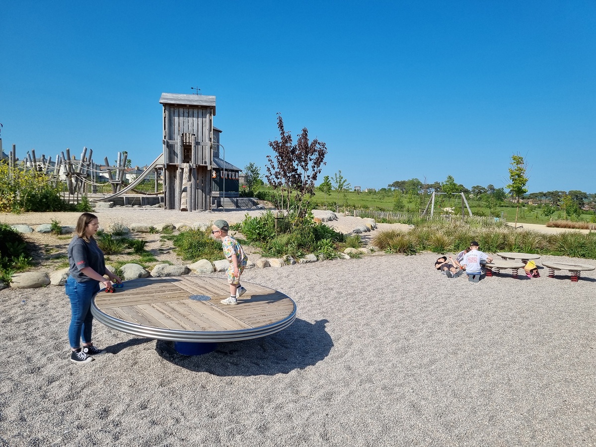 A mother and her child can be seen in the foreground. The child is standing on a rotating disc, the floor is covered with fine gravel. To the right, children are sitting in the gravel next to three jumping discs. In the background is a square tower.