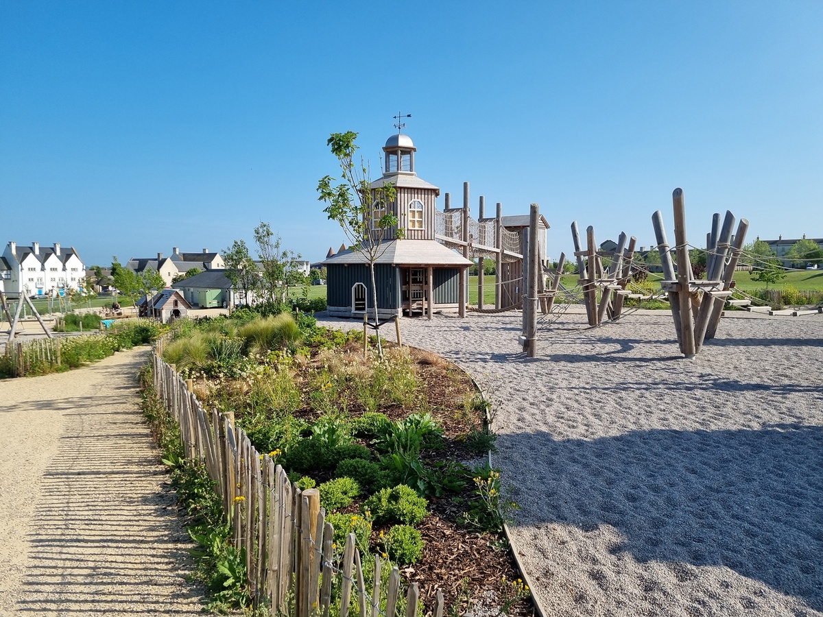 In the foreground is a flower bed with a wooden fence in front of it. To the left is a playground with fine gravel, triangular platforms and an angular wooden structure with windows and a weather cross on the roof. In the background are buildings.