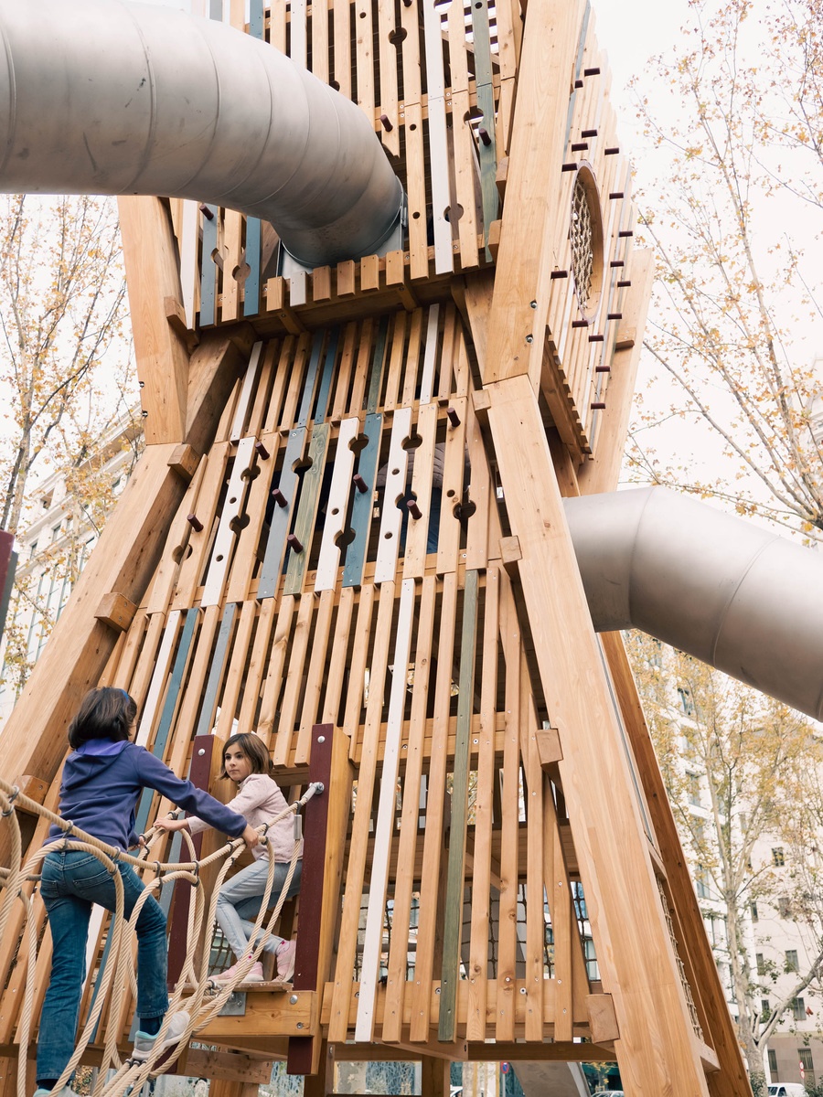 Zwei Mädchen klettern über eine Seilbrücke auf einen viereckigen Turm mit Tunnelrutschen. Der Turm besteht aus senkrechten Holzlatten mit ausgeschnittenen Formen und ist teilweise farbig bemalt. Im Hintergrund stehen Bäume mit herbstlich gefärbtem La