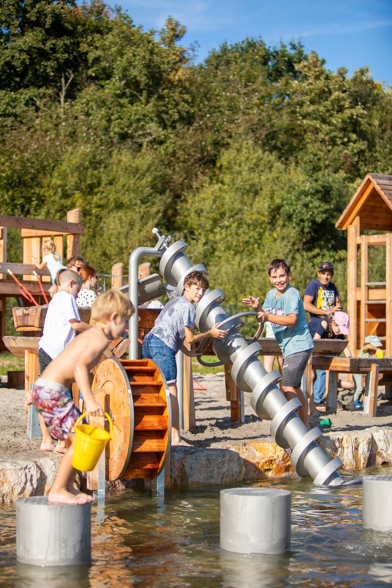 Kinder spielen mit verschiedenen Wasserspielgeräte, darunter Archimedischer Schraube und Mühlenrad am Bostalsee.