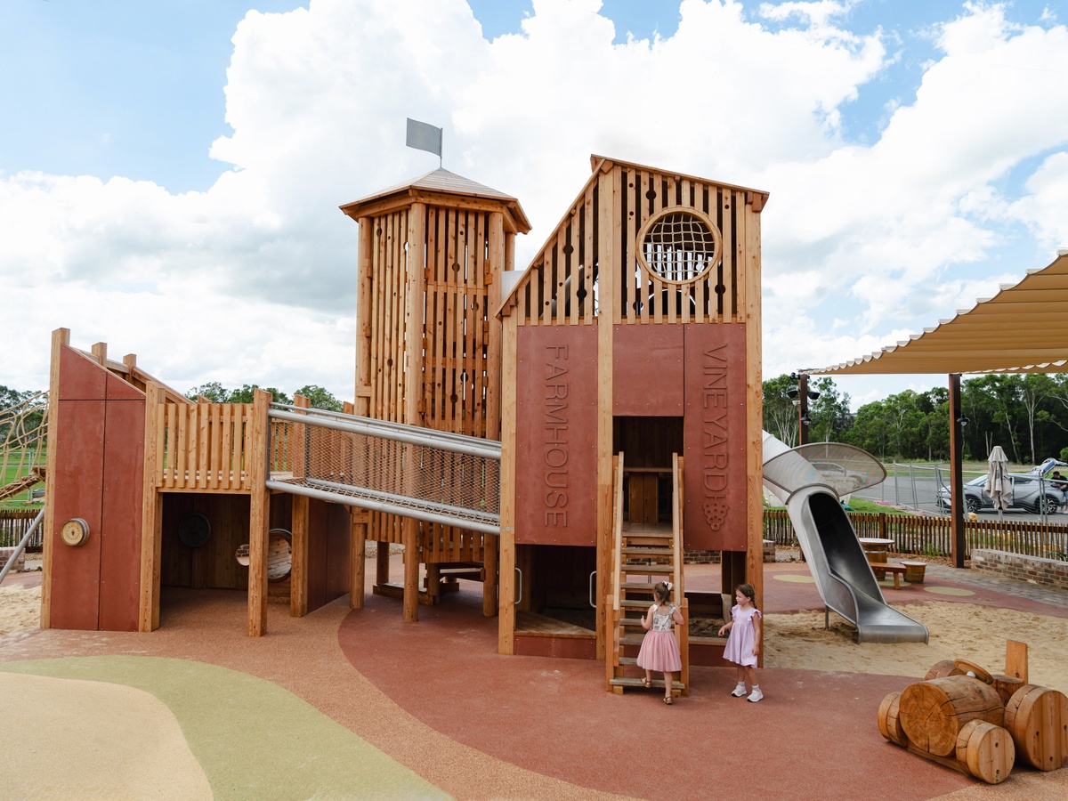 Spielanlage aus Holz mit der Aufschrift "Farmhouse Vineyard". Zwei Mädchen in rosa Kleidern stehen vor einer Holztreppe. Es gibt eine Tunnelrutsche, einen Brückenübergang, Kletterbereiche und einen Holztraktor. Im Hintergrund sind Bäume © Monique Sta