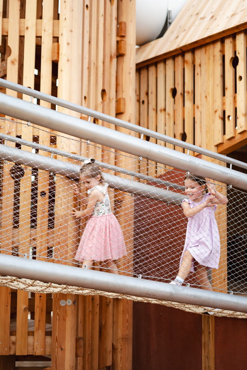 Zwei Mädchen auf Hängebrücke mit Metallgeländer, im Hintergrund Spielbereich mit ausgesparten Holzwänden © Monique Stano