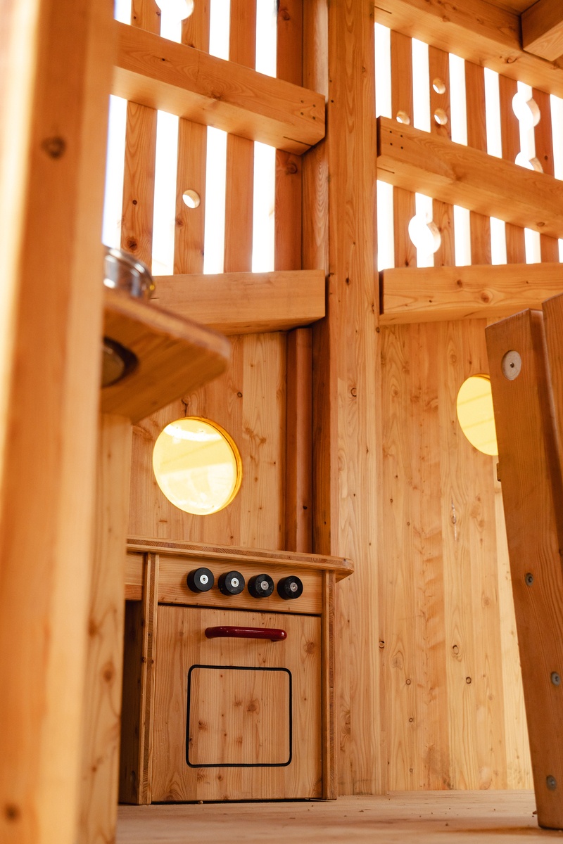 Innenraum des transparenten Turms mit vertikalen Holzlatten, zwei runden Fenstern und einer kleinen Spielküche mit Knöpfen und Ofentür. Das Design ist schlicht und rustikal mit natürlichen Holztönen © Monique Stano