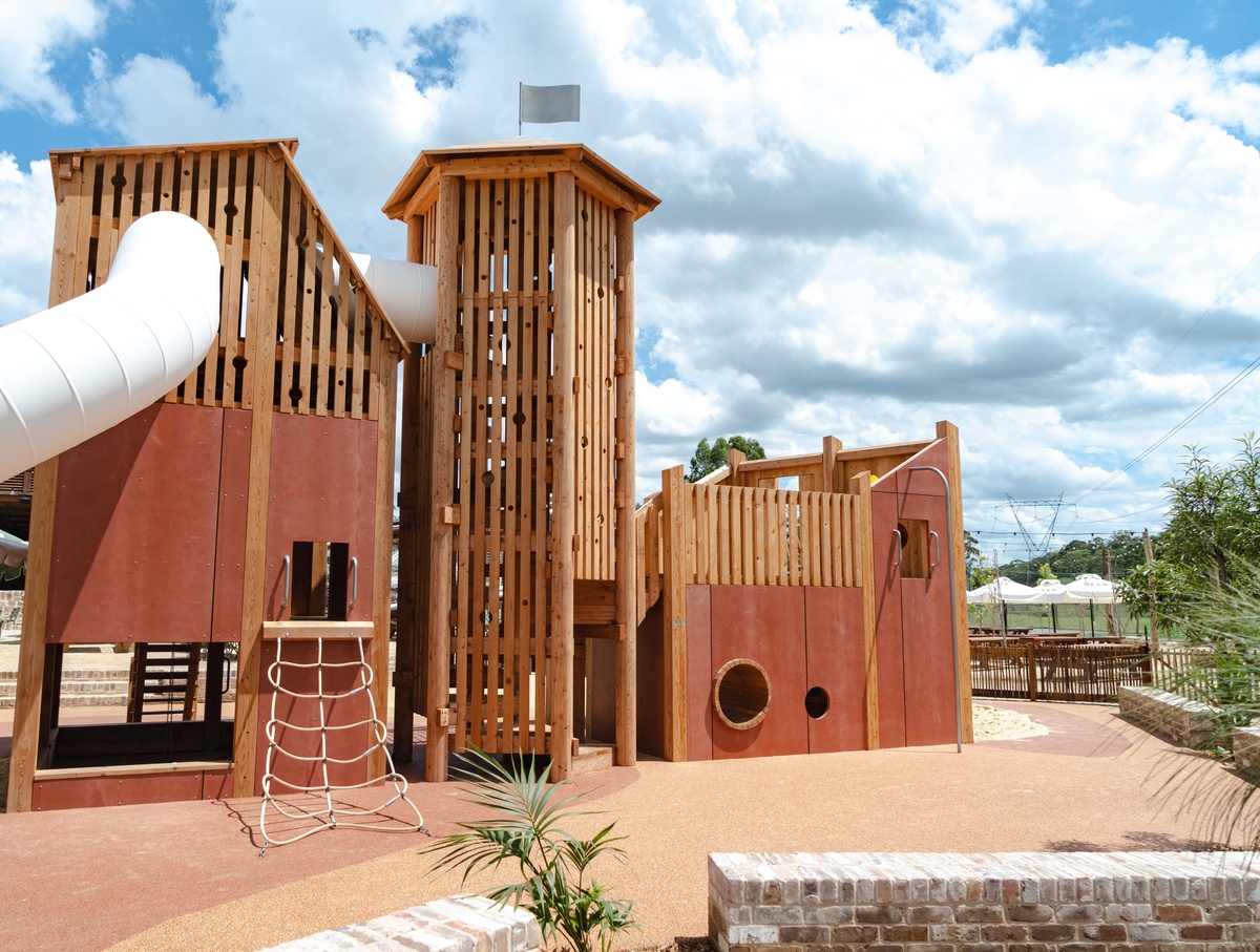 Wooden play area with tower and flag on the roof, sloping climbing net and tunnel slide, palm tree in front; partly cloudy sky © Monique Stano