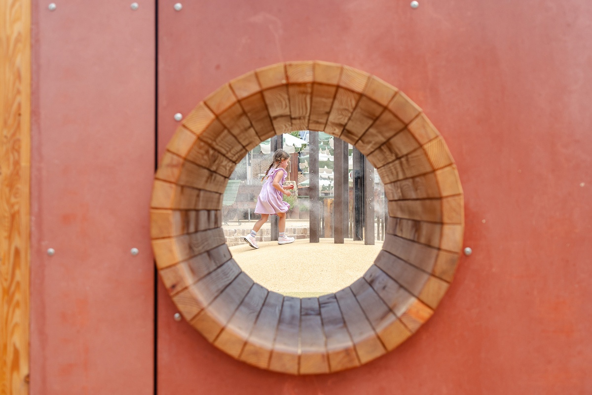 View trough wooden crawling barrel integrated into the wall, behind it a walking girl in a lilac-coloured dress on a sand-coloured fall protection surface © Monique Stano