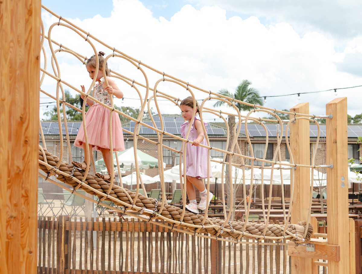 Two girls in light-coloured dresses on a rope bridge with PP rope crossing; in the background parasols, palm trees and roof with PV system © Monique Stano