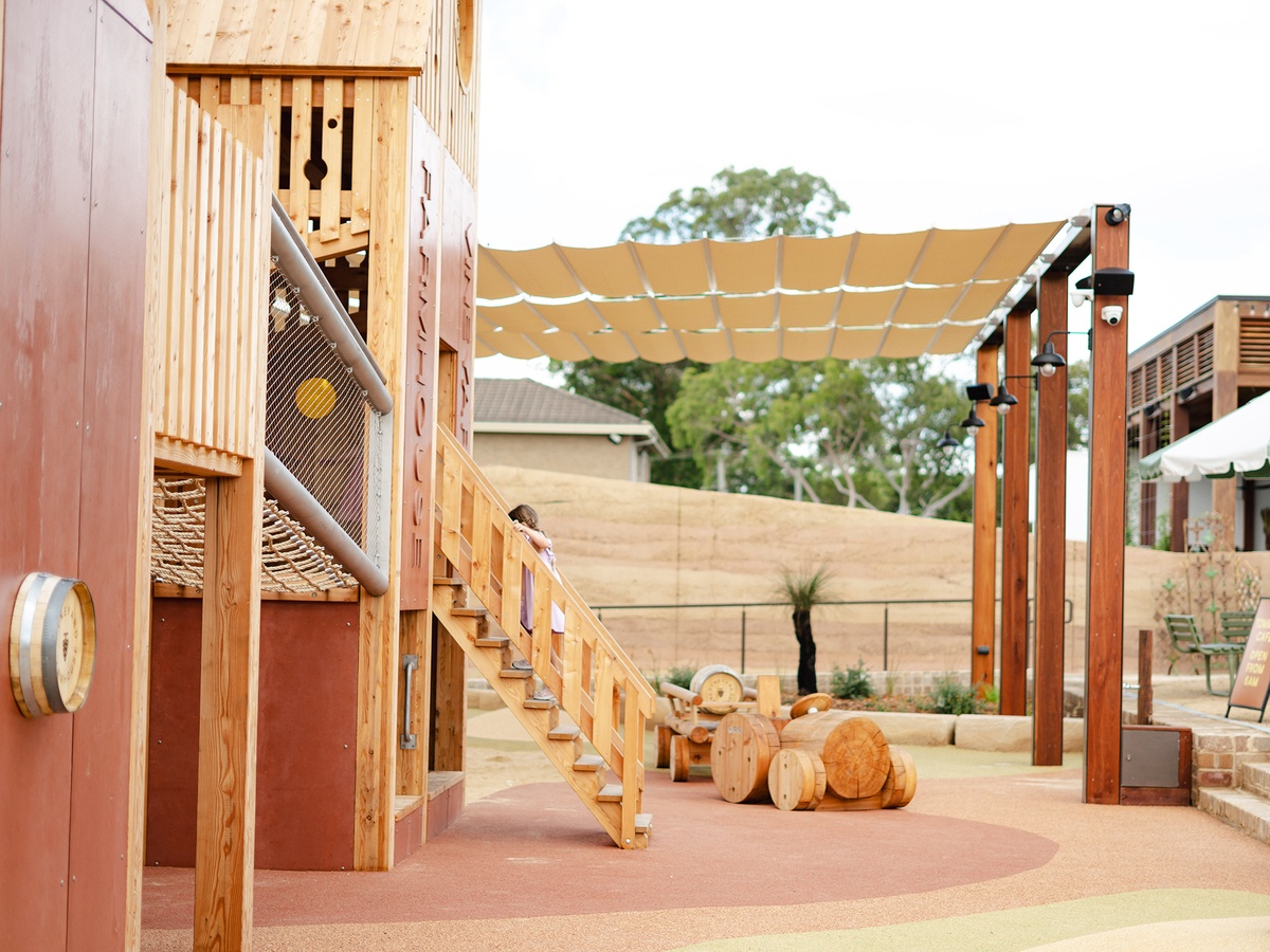 Play area with wooden tractor and trailer, rope bridge crossing with metal handrail and girl on wooden staircase. A sun sail, trees and buildings can be seen in the background © Monique Stano