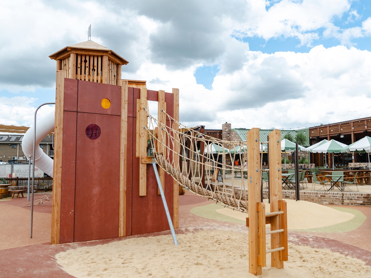 Unplayed play tower with flag on the roof, sliding pole, tunnel slide and suspension bridge with PP rope crossing. Behind it, white and mint green parasols and buildings and a partly cloudy sky © Monique Stano