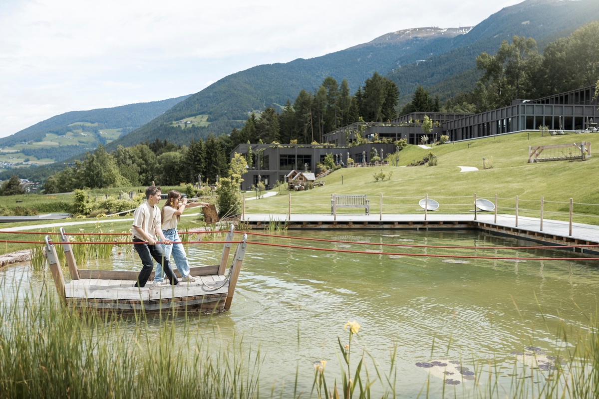 Zwei Kinder stehen auf einer Seilfähre im Wasser, im Hintergrund sind Berge und eine Spiellandschaft auf einer Wiese zu sehen © Alex Filz