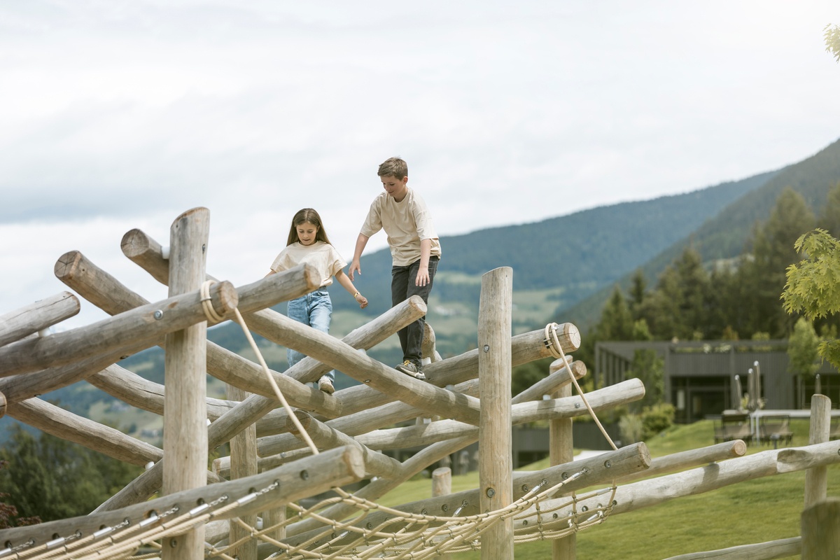 Ein Junge und ein Mädchen klettern auf der Kletterstruktur 16.  Im Hintergrund sind Bäume, Gebäude und Berge zu sehen. Das Wetter ist heiter. © Alex Filz