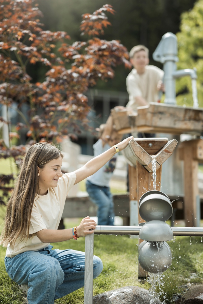 Ein Mädchen steht neben einem kleinen Schaufelrad, auf das Wasser von einer Holzrinne spritzt. Im Hintergrund steht ein Junge an einer Spielplatzpumpe und pumpt Wasser in eine Holzrinne. Das Wetter ist heiter. © Alex Filz