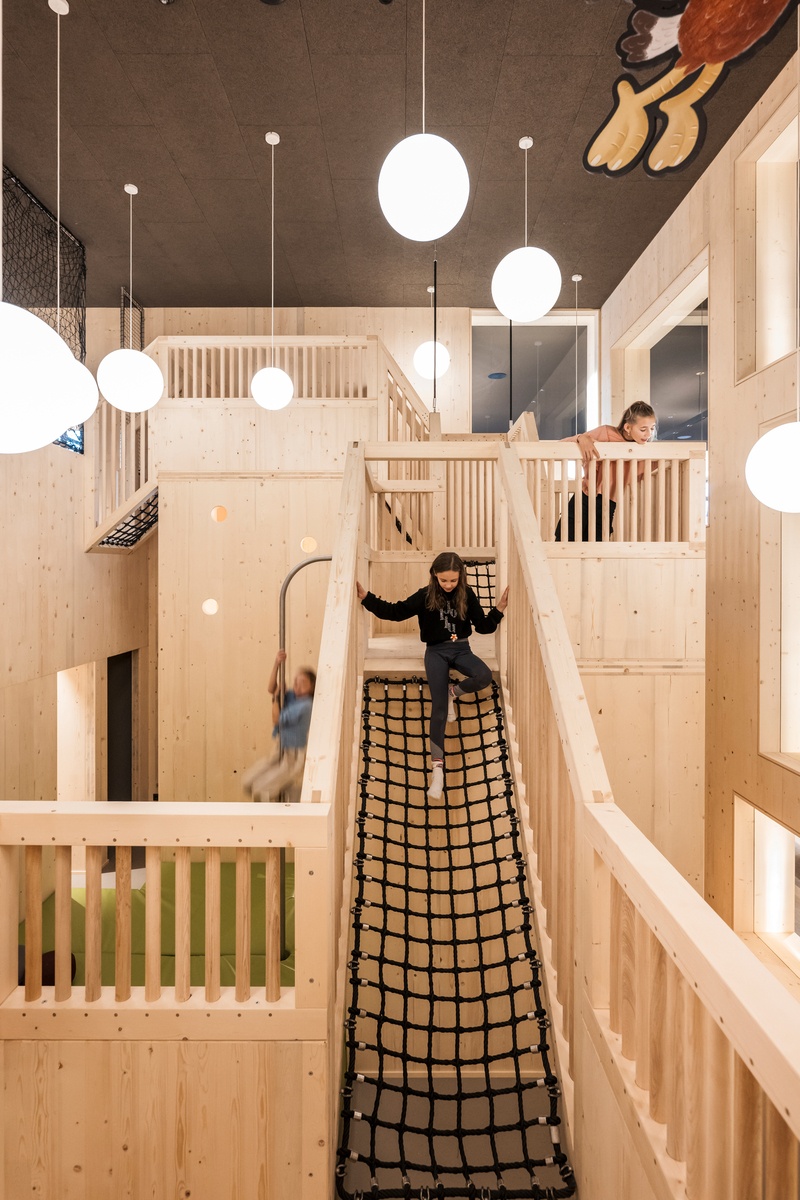 Kinder spielen in einem hölzernen Indoor Spielbereich mit Netzübergängen. Ein Kind rutscht eine gebogene Rutschstange hinab. Von der Decke hängen weiße kugelartige Leuchten © Alex Filz